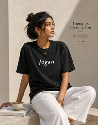 Woman wearing a black 'Jogan' t-shirt sitting with a cup, against a neutral background.